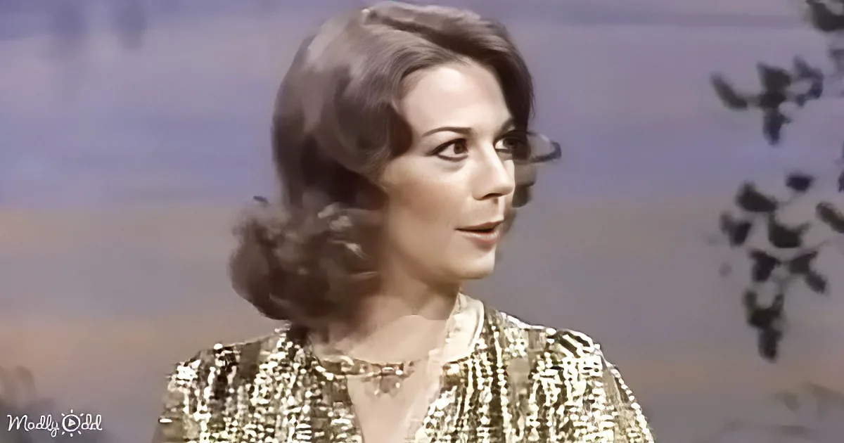 A woman with dark, styled hair wearing a patterned top and necklace, set against a blurred background.