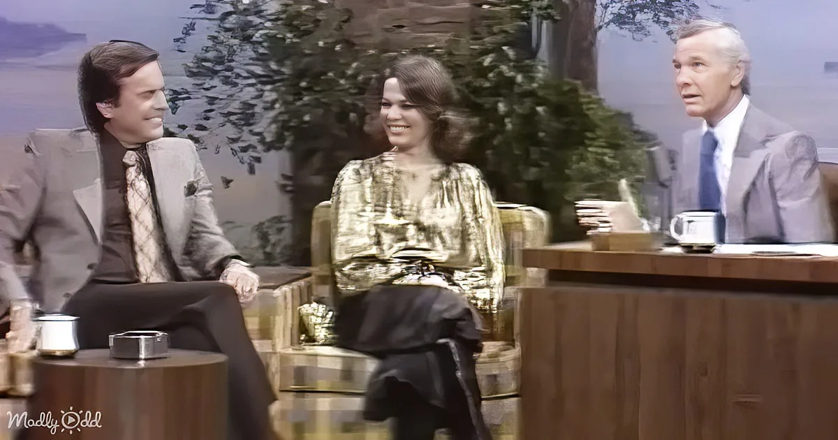 A woman in a gold dress sits between two men in suits on a talk show set.