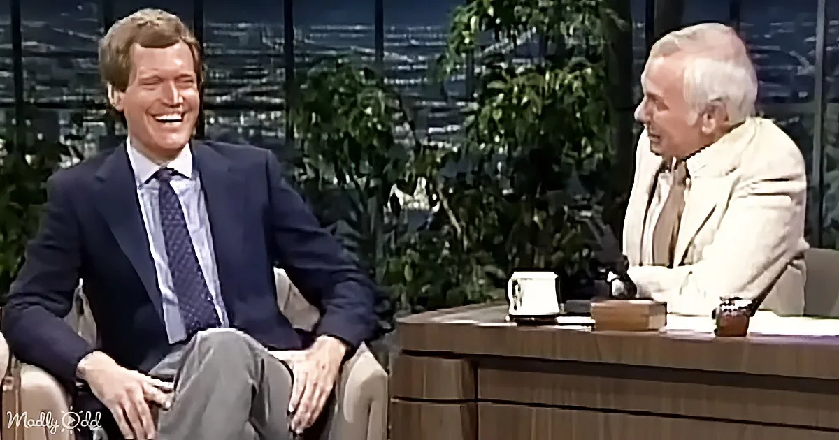 A man in a suit sits on a couch, laughing, while another man in a light-colored suit sits at a desk, also laughing.