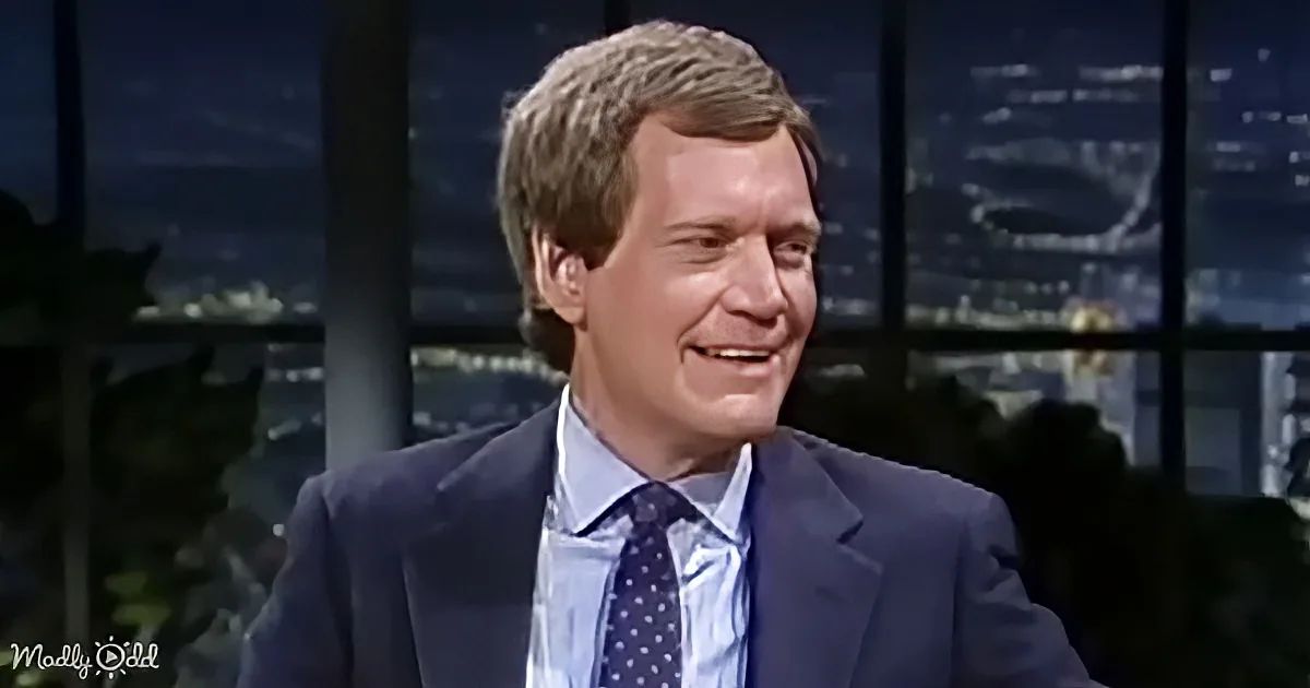 A man in a suit and tie smiles while sitting in front of a cityscape backdrop.