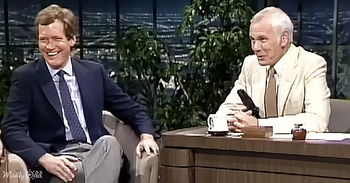 Two men in suits, one laughing, seated on a talk show set with a cityscape background.