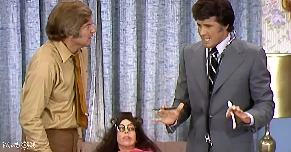 A man in a suit gestures emphatically while another man in a beige shirt looks on, with a woman in glasses reclining on a...