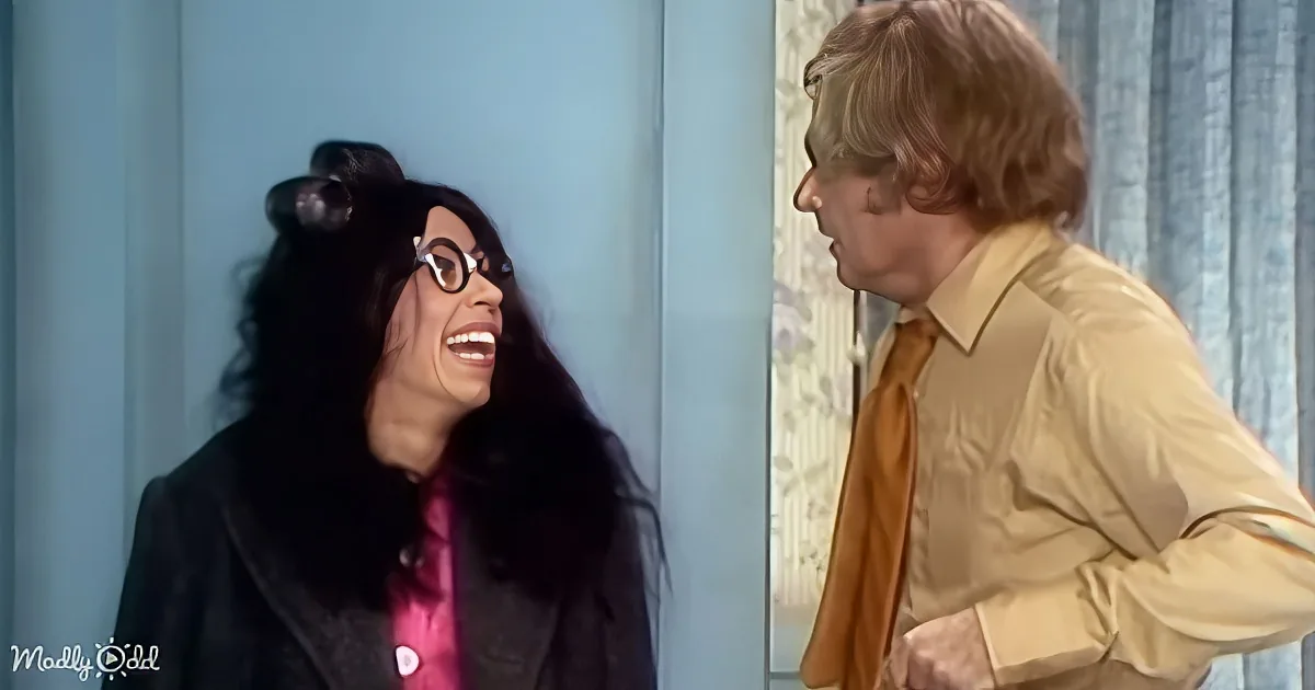 A woman with long black hair and glasses laughs while a man in a yellow shirt and orange tie looks at her.