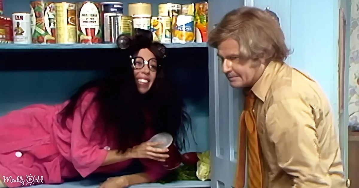 A woman in a pink robe smiles while holding a container, with a man in a yellow shirt looking on.
