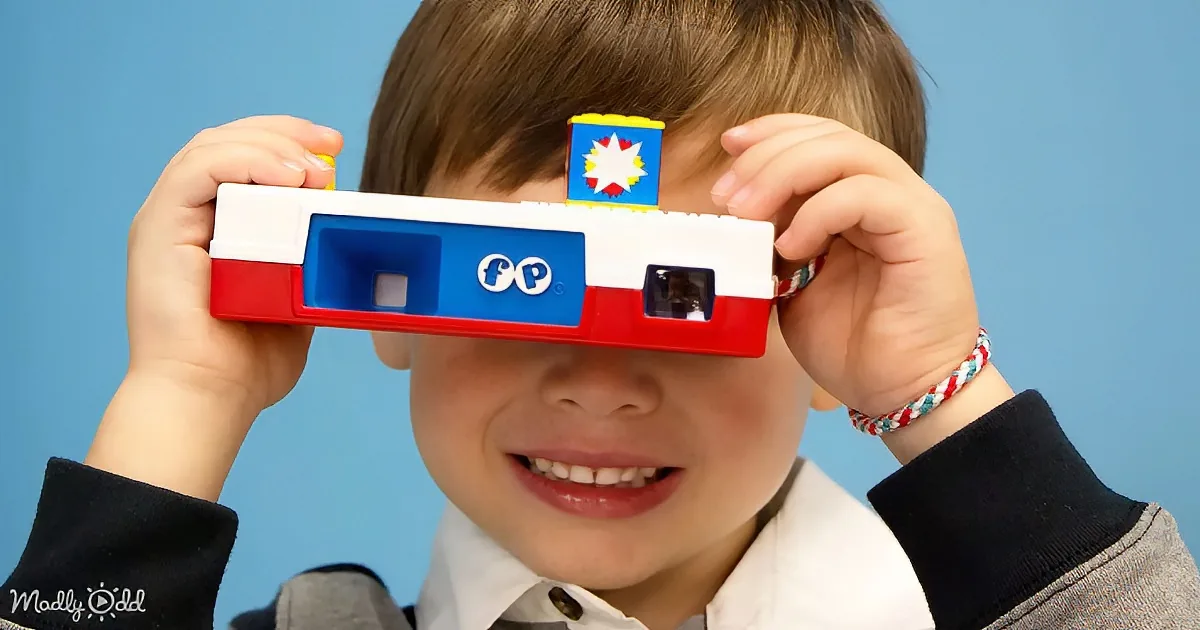 A child holds a red, white, and blue toy camera with a "FP" logo, smiling against a blue background.