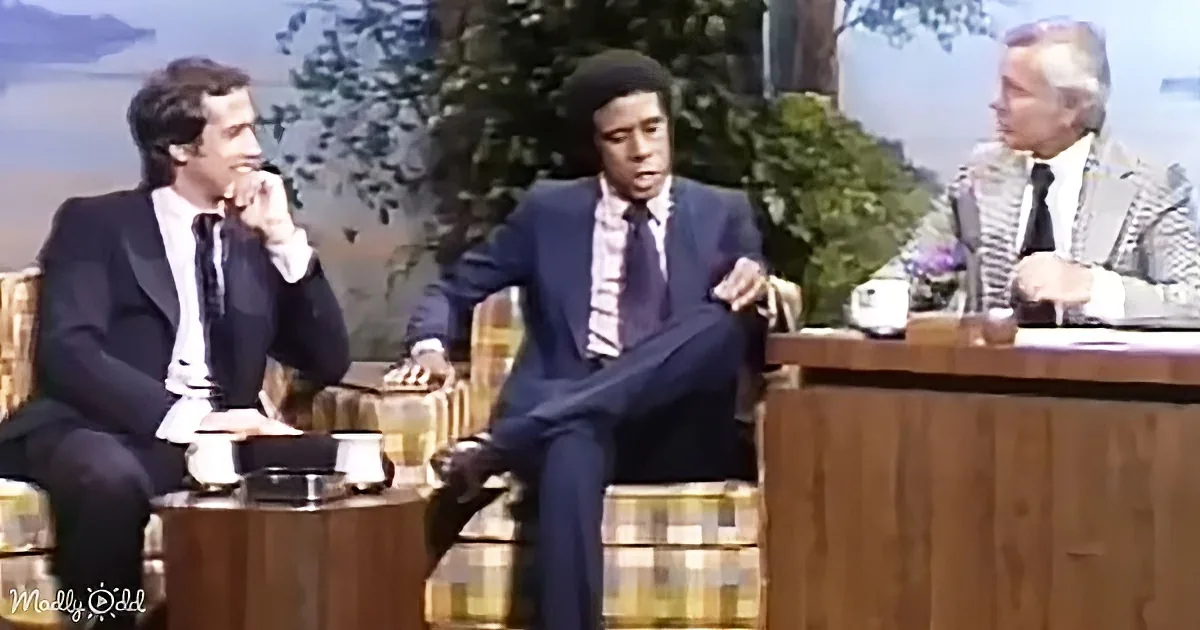 A man in a suit and tie is sitting on a couch, gesturing with his hands while talking.