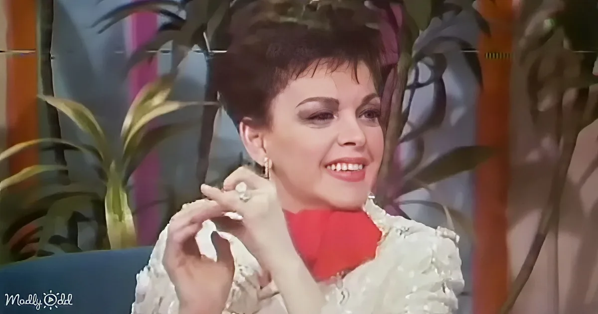A woman with short hair, wearing a white lace top and red scarf, smiles while holding a piece of food.