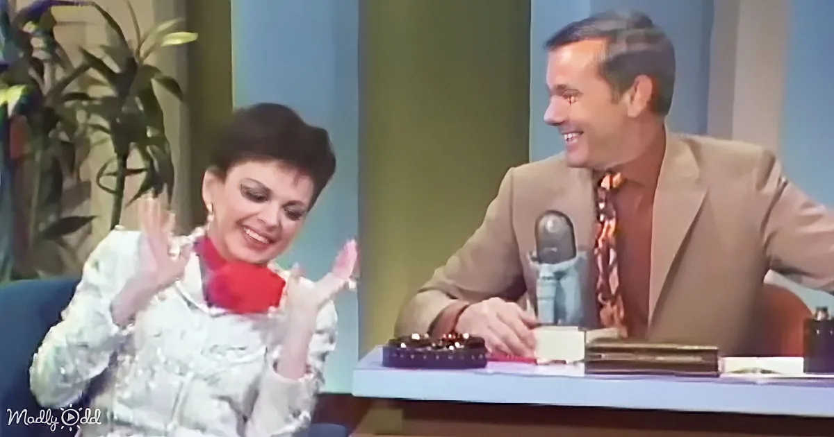 A woman in a white outfit and red scarf gestures animatedly while seated next to a man in a beige suit and patterned tie.