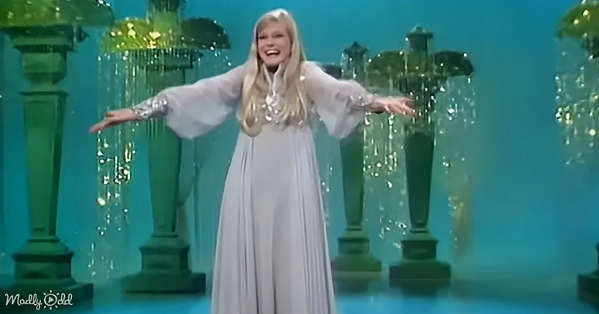 A woman in a flowing white gown with arms outstretched stands on a stage with green pillars and sparkling water effects.