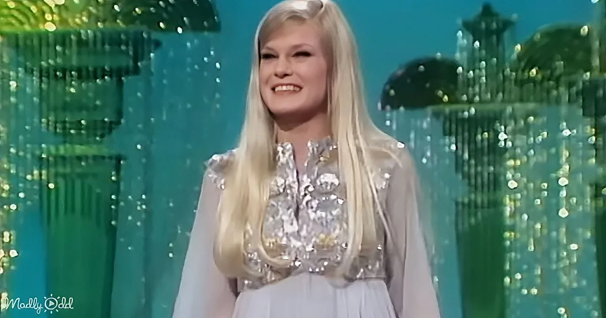 A woman with long blonde hair smiles in a white dress with sequins, standing in front of a glittery green background.