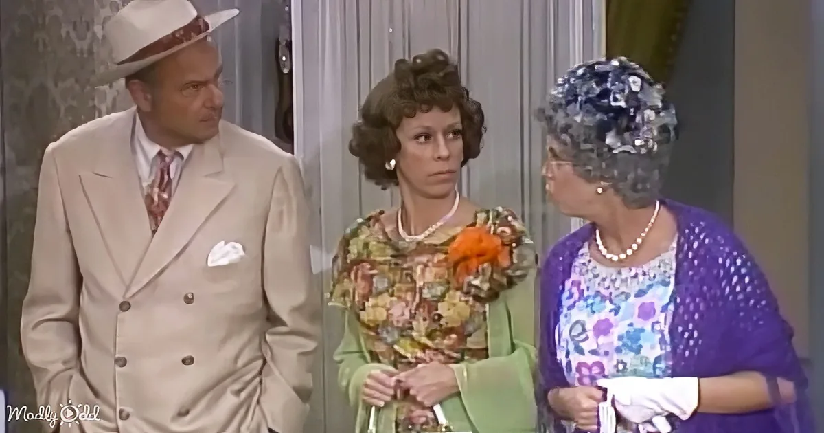 A man in a beige suit and hat stands beside two women, one in a floral dress and the other in a purple outfit with a...