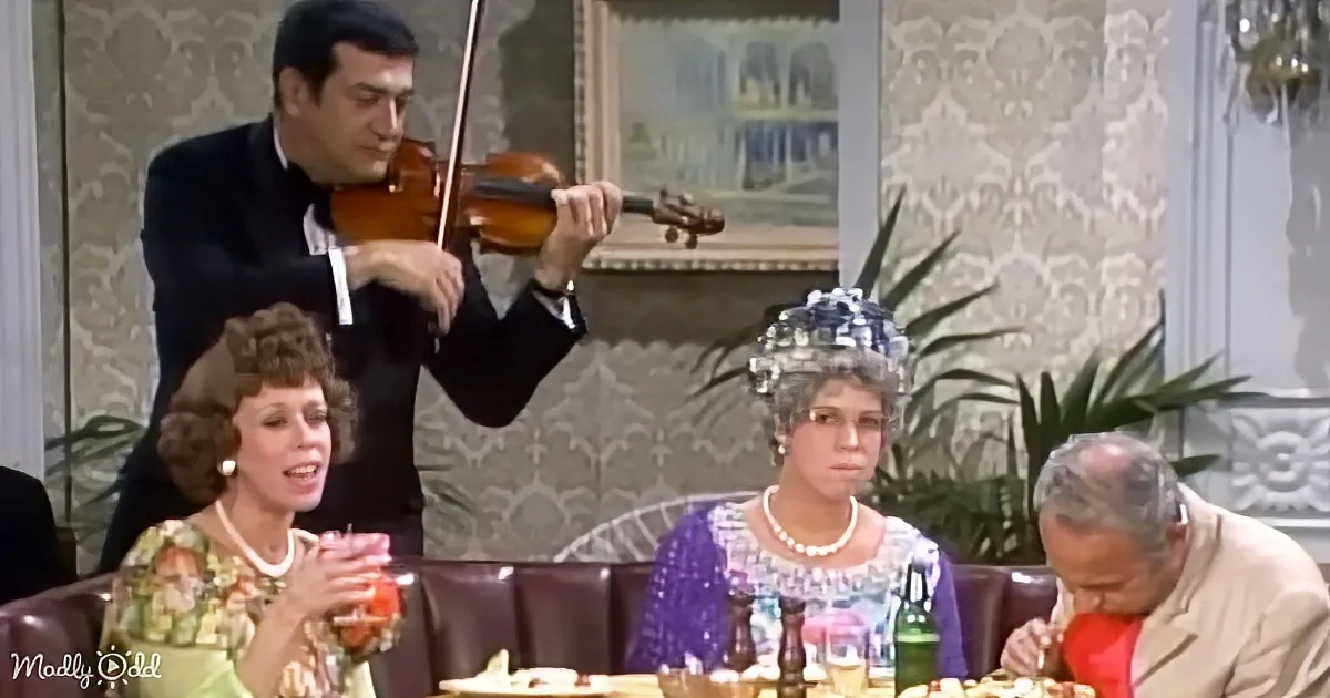 A man in a tuxedo plays the violin while a woman in a floral dress and another in a purple dress sit at a table...