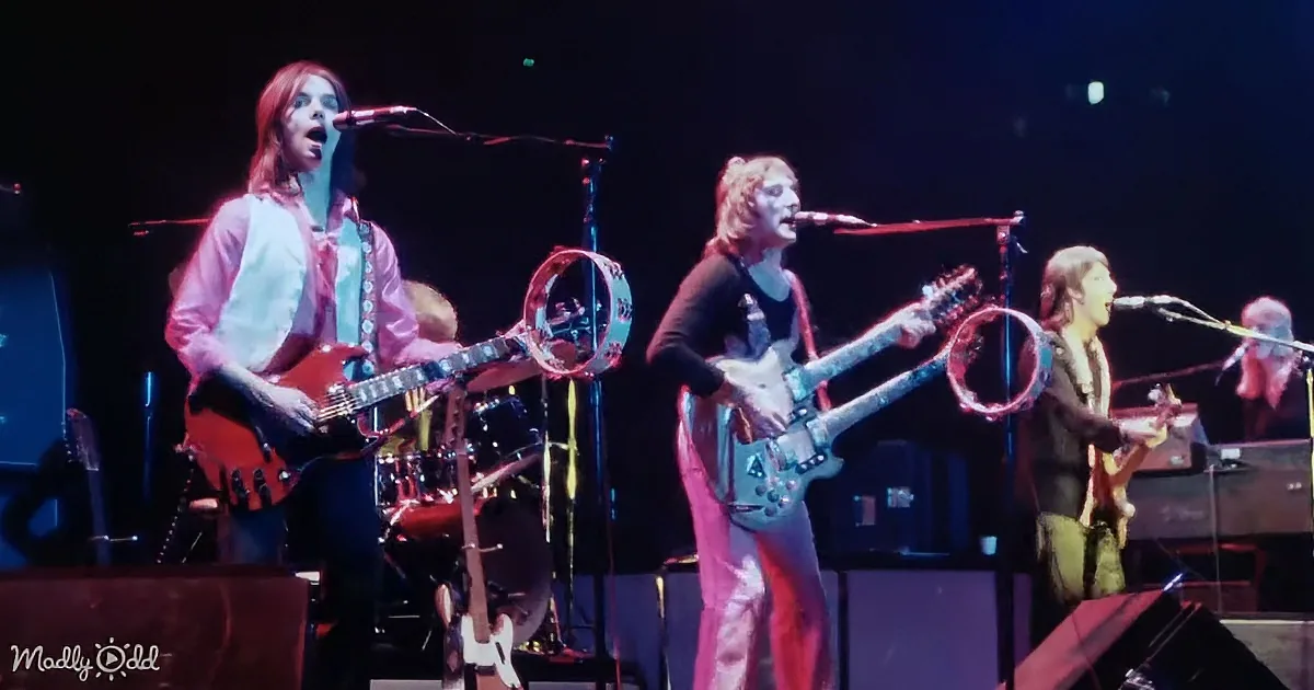A band performs on stage with two guitarists singing, one in pink and another in black, surrounded by musical equipment.