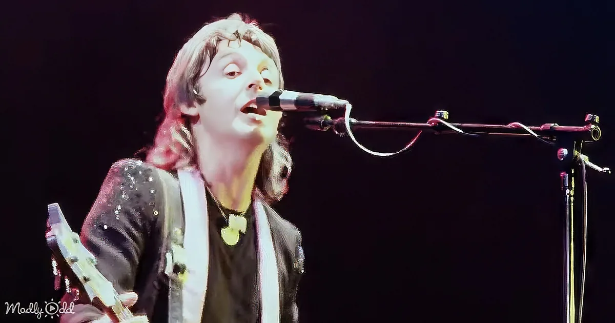 A person singing into a microphone while holding a guitar, wearing a black jacket with sequins and a necklace.