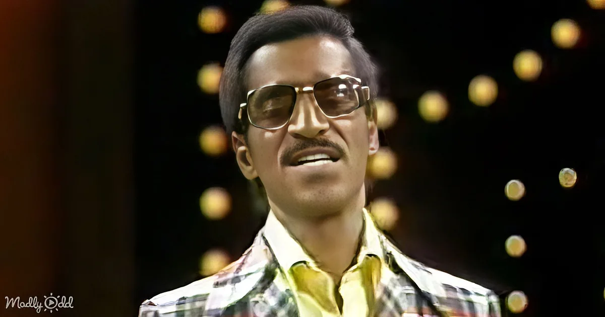 A man in a patterned shirt and sunglasses, smiling, with a blurred background of lights.