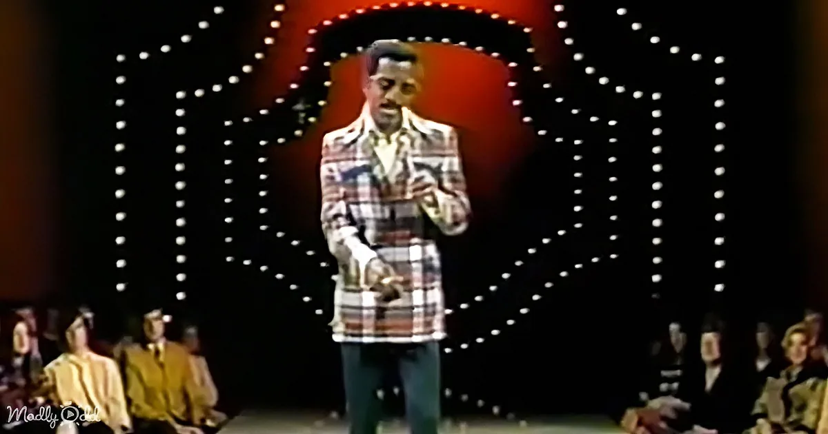 A man in a plaid suit stands on stage with a microphone, surrounded by an audience and a circular light pattern.