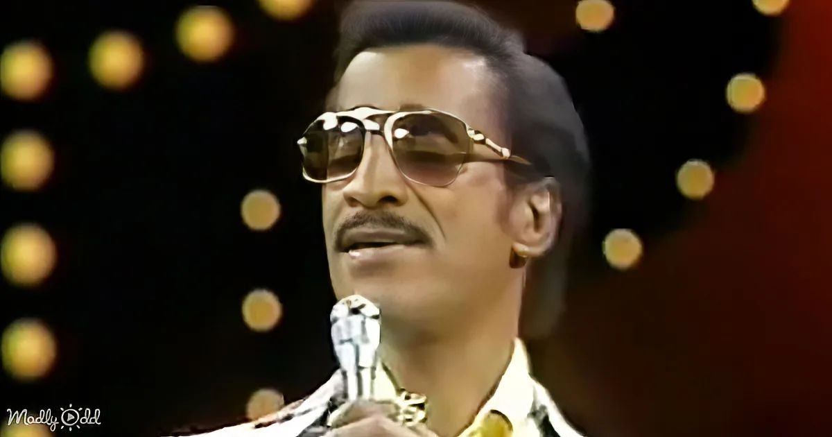 A man wearing sunglasses and a patterned shirt is holding a microphone, with a bokeh effect in the background.