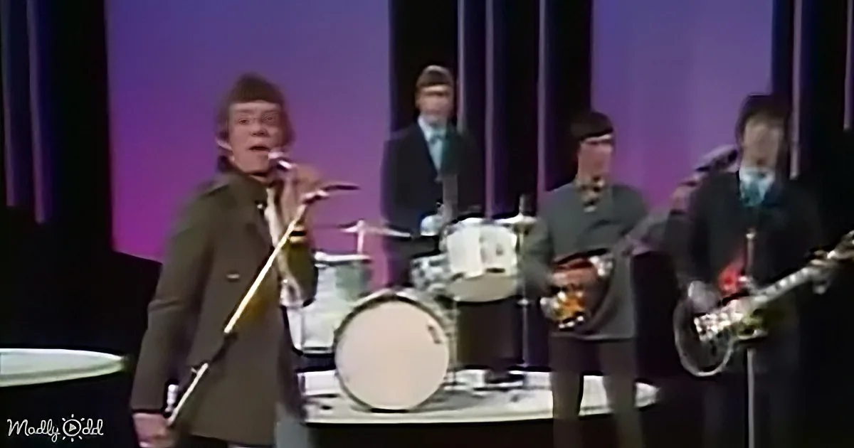 A band member in a green jacket sings into a microphone on stage with other musicians.