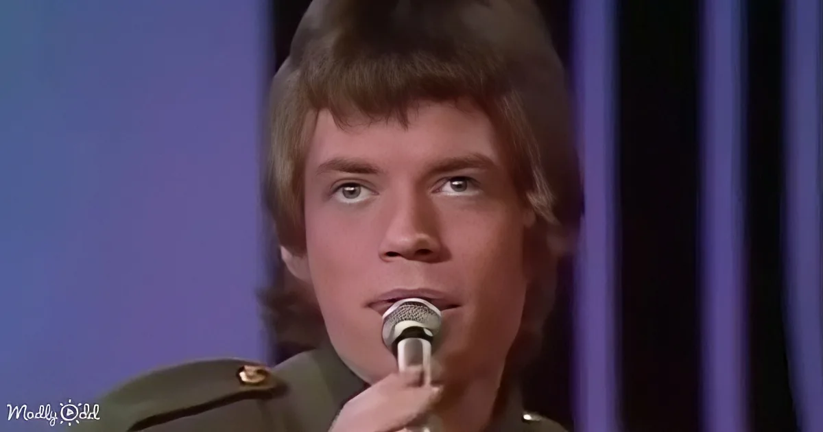 A person in a military-style uniform holds a microphone, with a purple background.