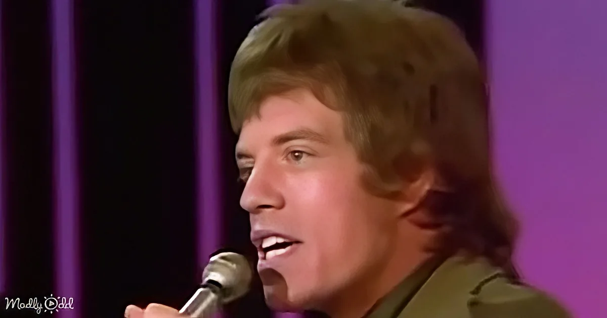 A person with shoulder-length hair sings into a microphone against a purple background.