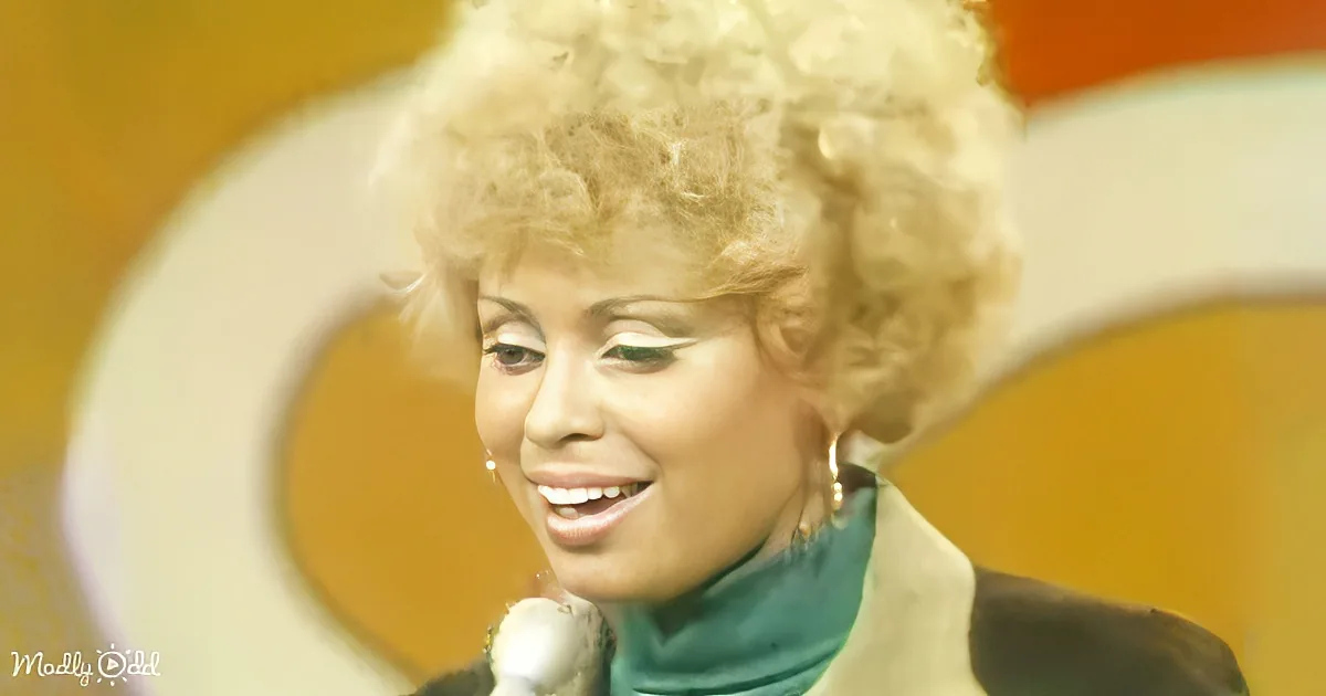 A smiling woman with curly blonde hair, wearing a green turtleneck and beige jacket, holding a microphone.