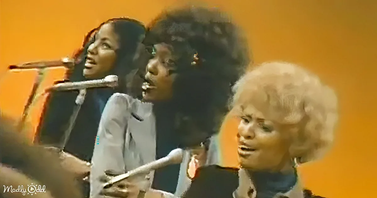 Three women singing into microphones, one with a large afro, against an orange background.