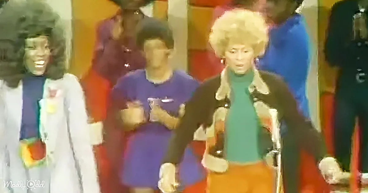 A woman with a large afro hairstyle stands in front of a group of people, holding a bottle.