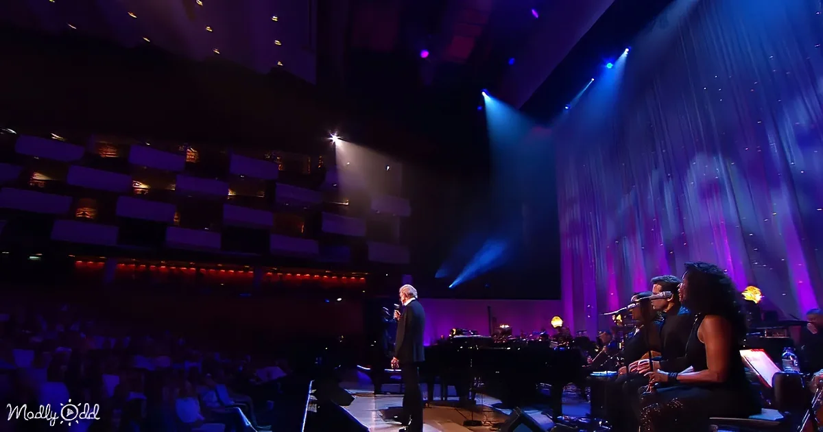 A man in a suit sings on stage with a band and microphone, under colorful stage lights.