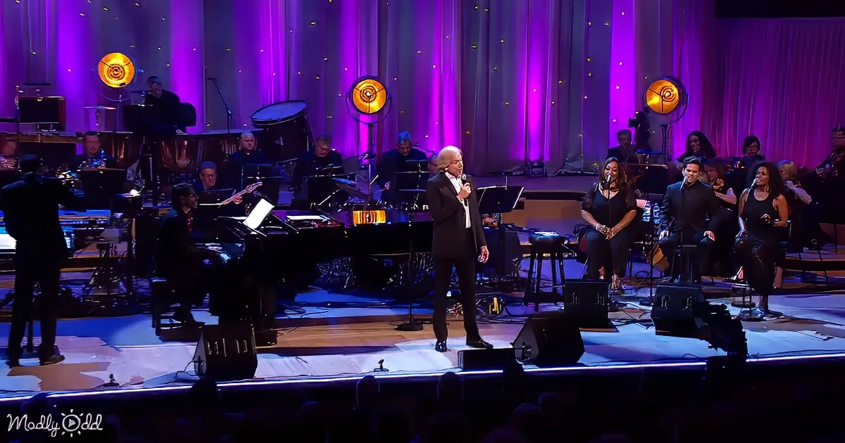 A man in a suit sings on stage with a band and orchestra behind him.