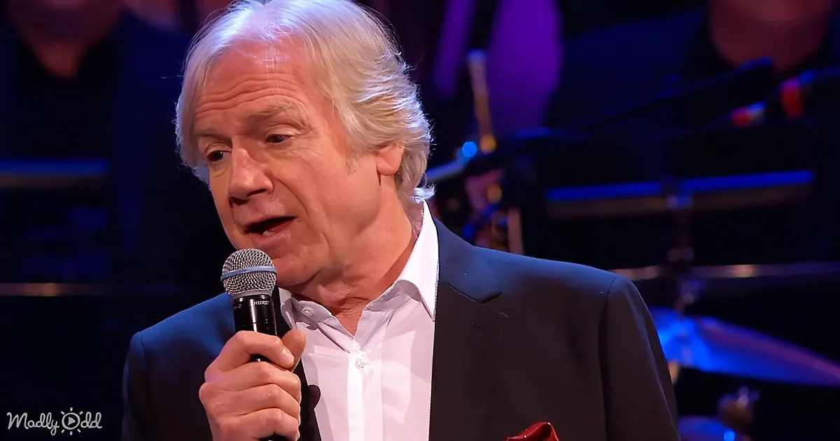 An older man with white hair, wearing a suit and holding a microphone, appears to be speaking or singing on stage.