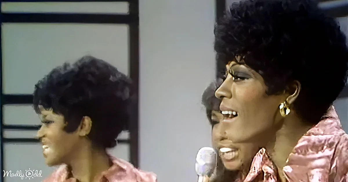 A woman with an afro hairstyle sings into a microphone, wearing a pink outfit and gold earrings.
