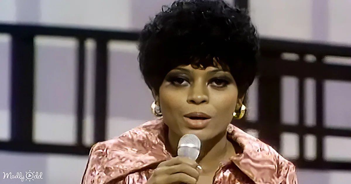 A woman with curly hair, wearing a pink outfit, holds a microphone and appears to be speaking or singing.