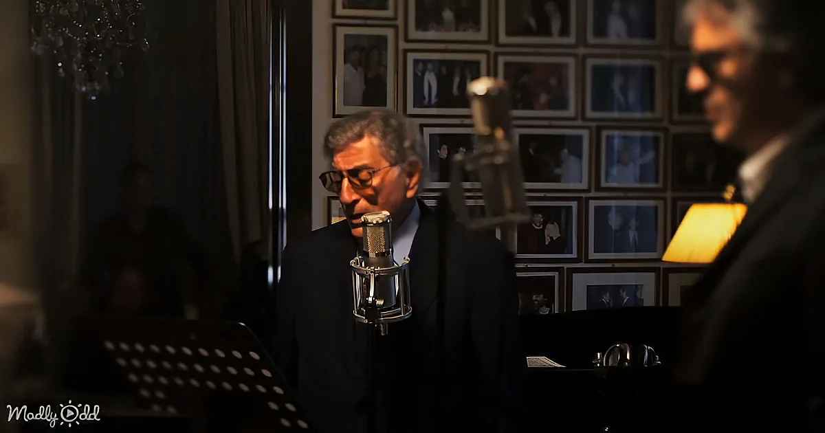 A man in a suit sings into a microphone in a dimly lit room with framed photos on the wall.