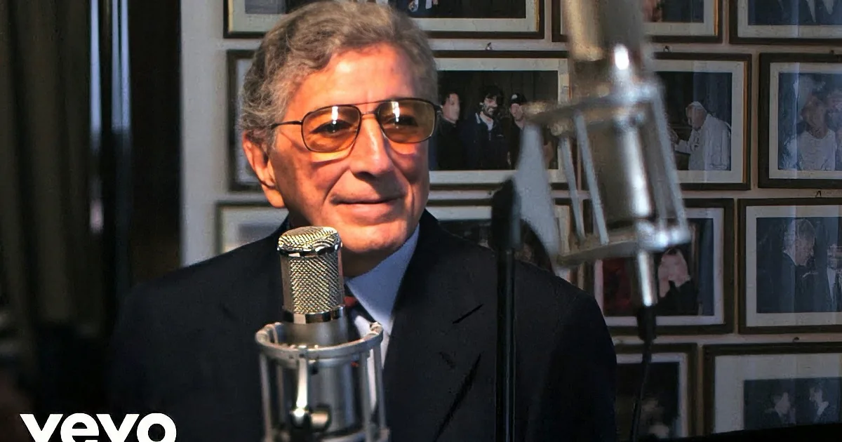 A man in a suit and sunglasses stands in front of a microphone, facing another man in a dimly lit room with framed...