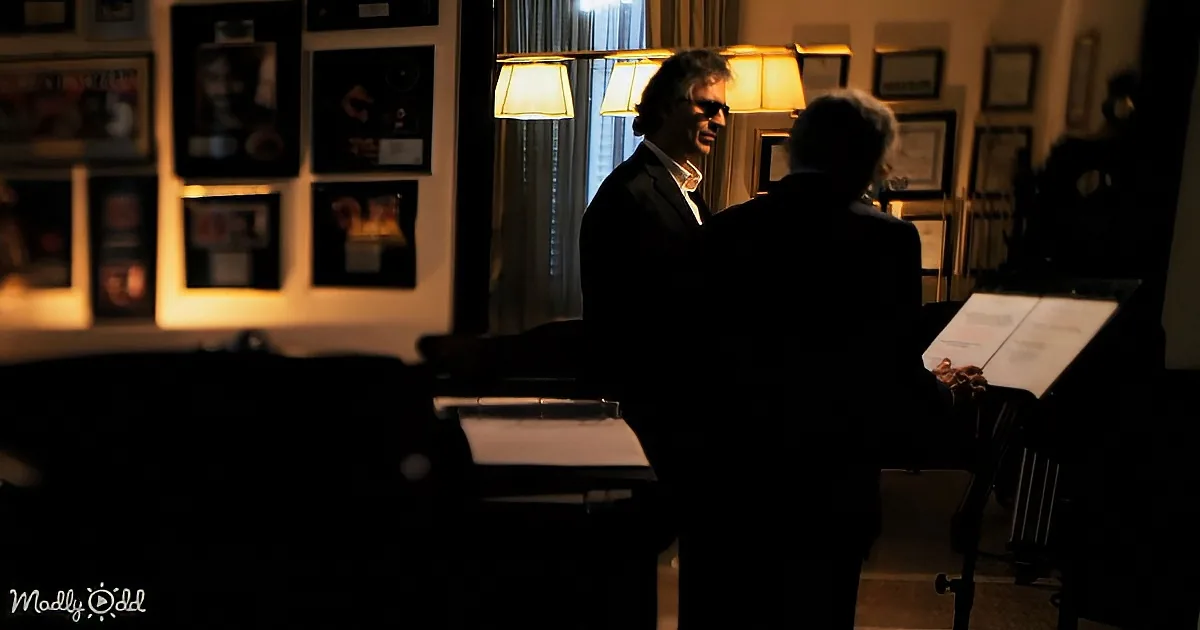 Two men in a dimly lit room, one holding sheet music, another in a suit, surrounded by framed pictures and lamps.
