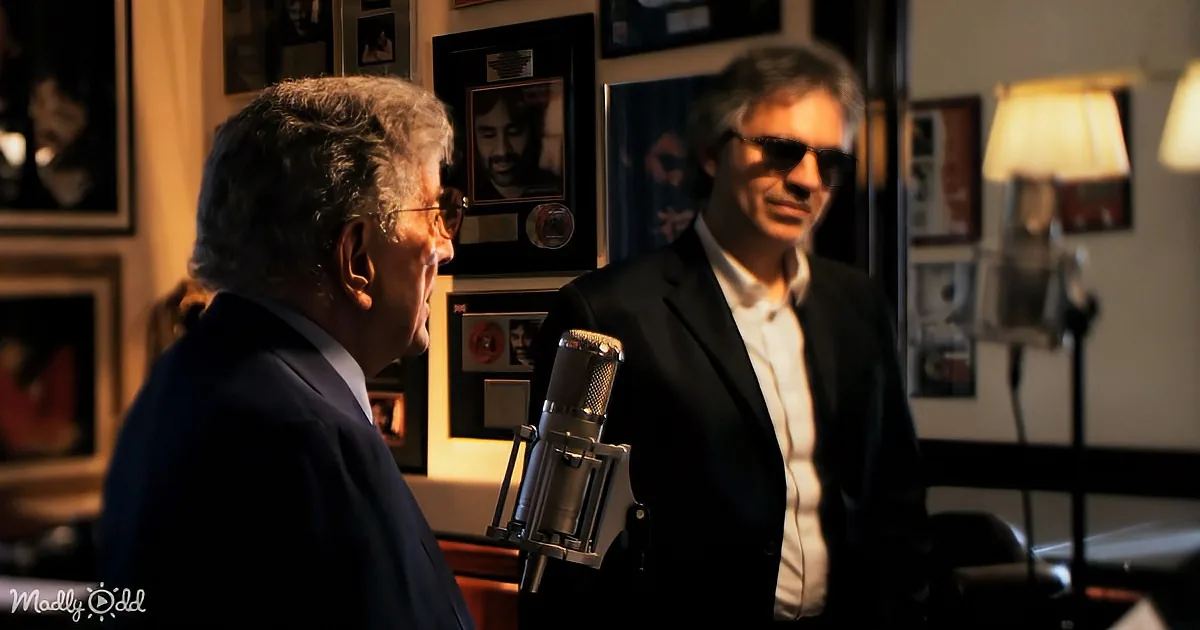 Two men in a recording studio, one speaking into a microphone, surrounded by framed photos and awards.