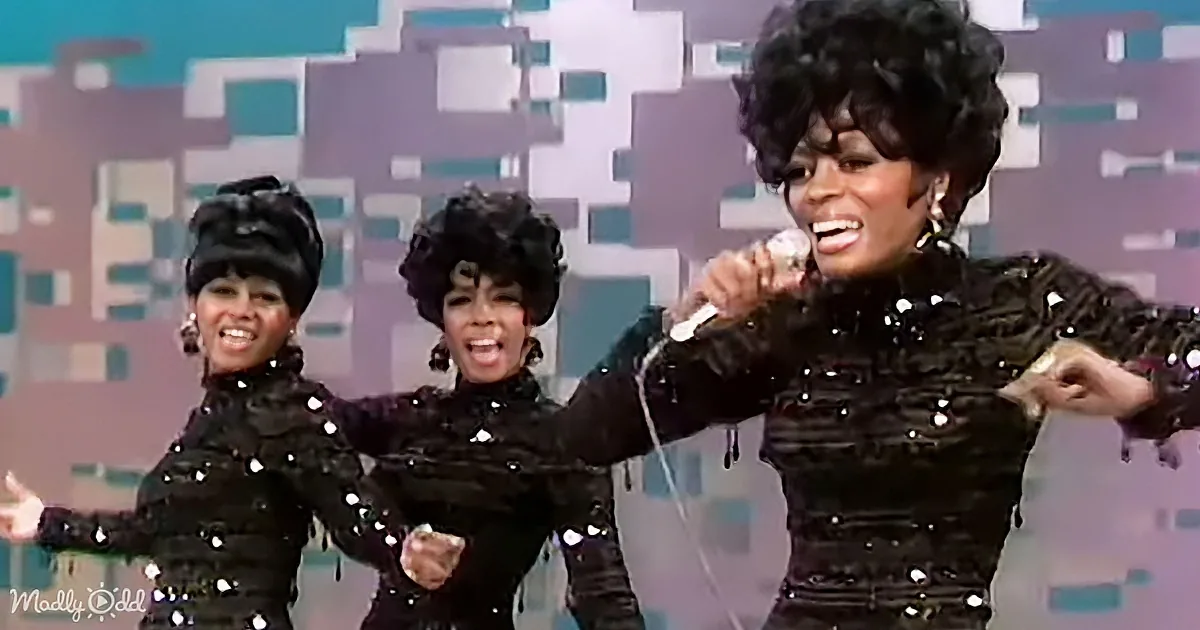 A woman in a black sequined outfit sings into a microphone, accompanied by two dancers in matching outfits.