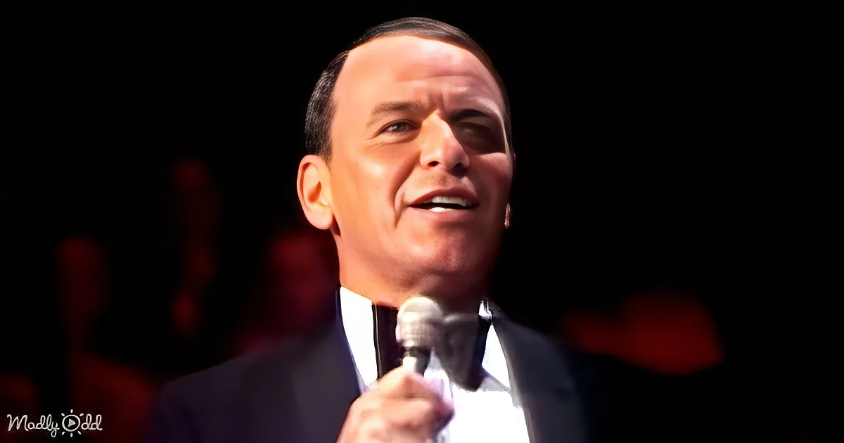 A man in a suit holds a microphone, smiling, with a dark background and red lighting.