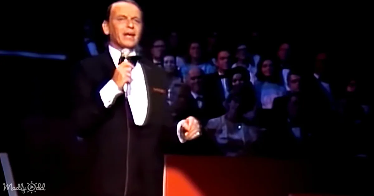 A man in a tuxedo sings into a microphone on stage, with an audience in the background.