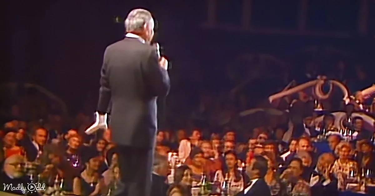 A man in a suit stands on stage, facing an audience, holding a microphone.