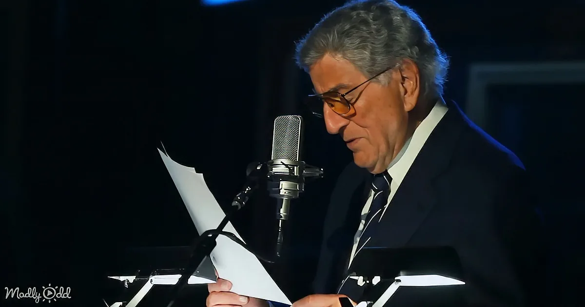 A man in a suit and glasses reads from a paper into a microphone on stage.