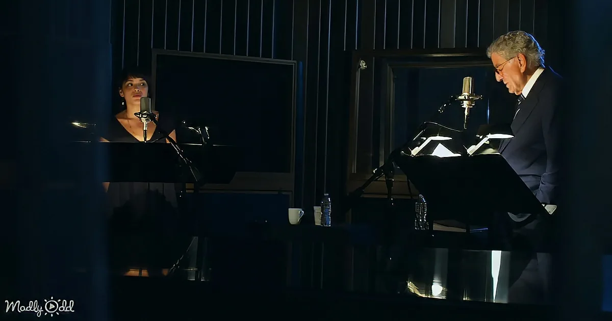 A man in a suit sits at a piano with a microphone, while a woman stands behind him, also with a microphone.