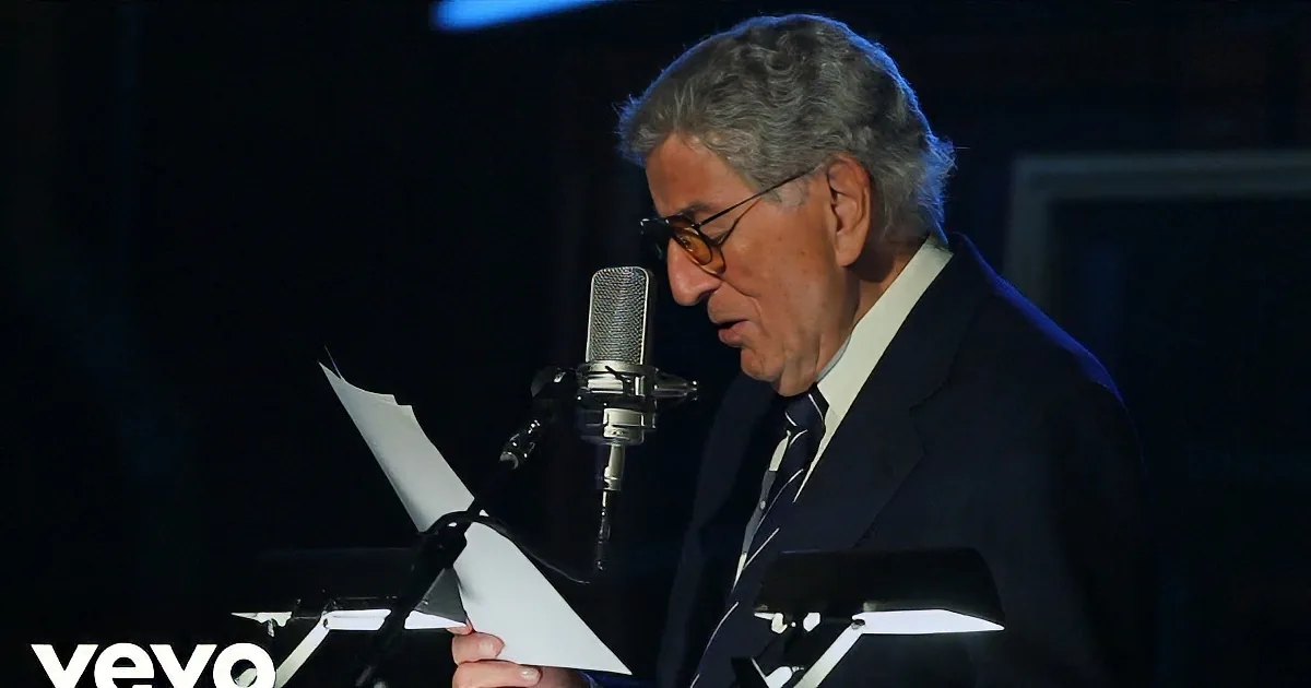 An older man in a suit sings into a microphone, with a woman in a smaller inset screen singing behind him.