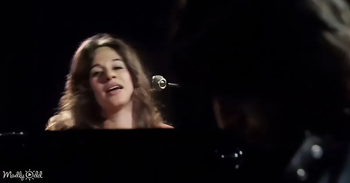A woman with long hair sings into a microphone, illuminated against a dark background.