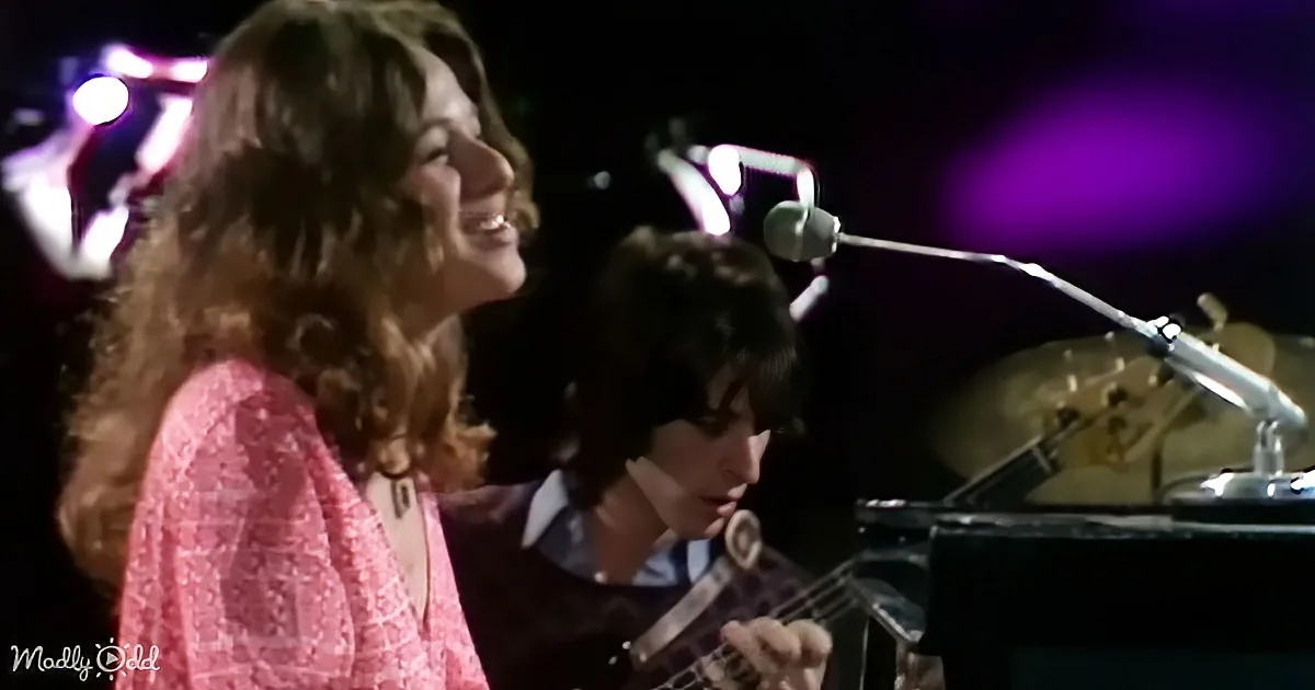 A woman with long hair smiles while a man plays guitar in a dimly lit setting with purple lights.