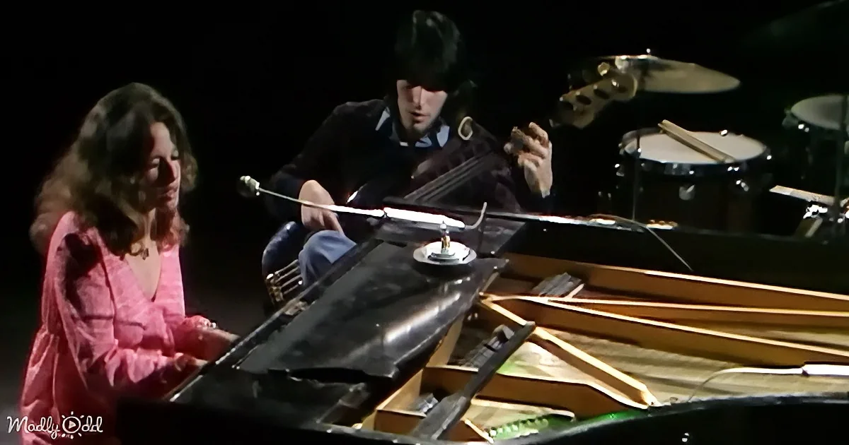 A woman in pink plays piano while a man in dark clothing plays a stringed instrument.