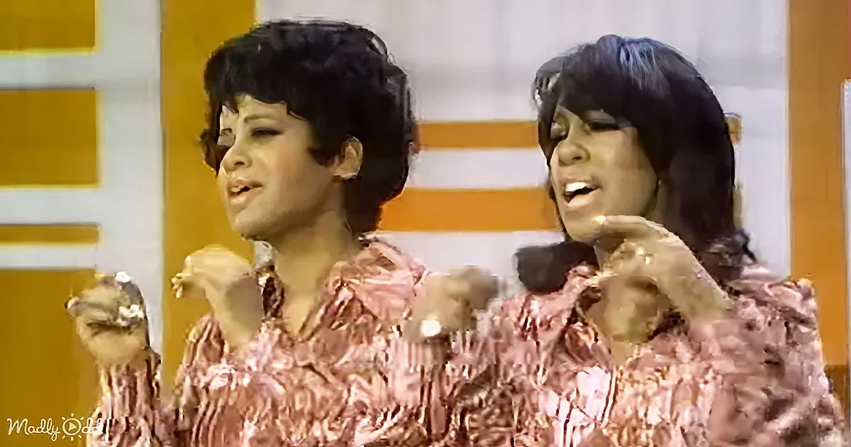 Two women in matching pink floral shirts are singing and gesturing with their hands.