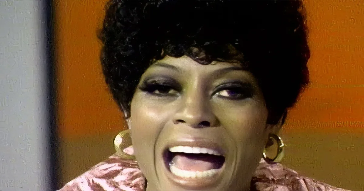 A woman with short, curly hair, wearing gold hoop earrings and a pink top, smiles softly against an orange background.