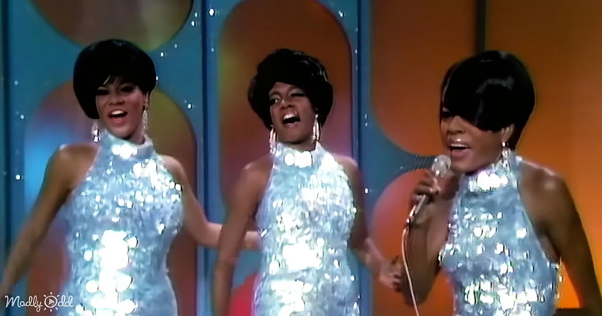 Three women in shimmering silver dresses perform on stage with a colorful backdrop.
