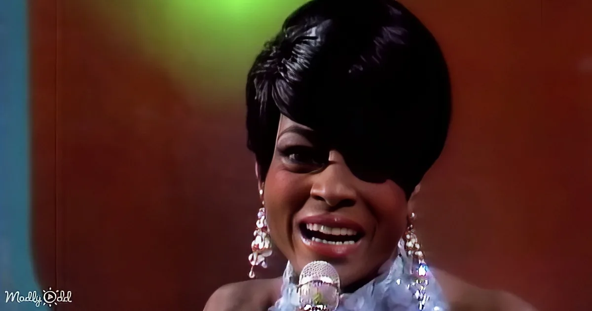 A woman with short black hair and large earrings sings into a microphone, wearing a light blue outfit.